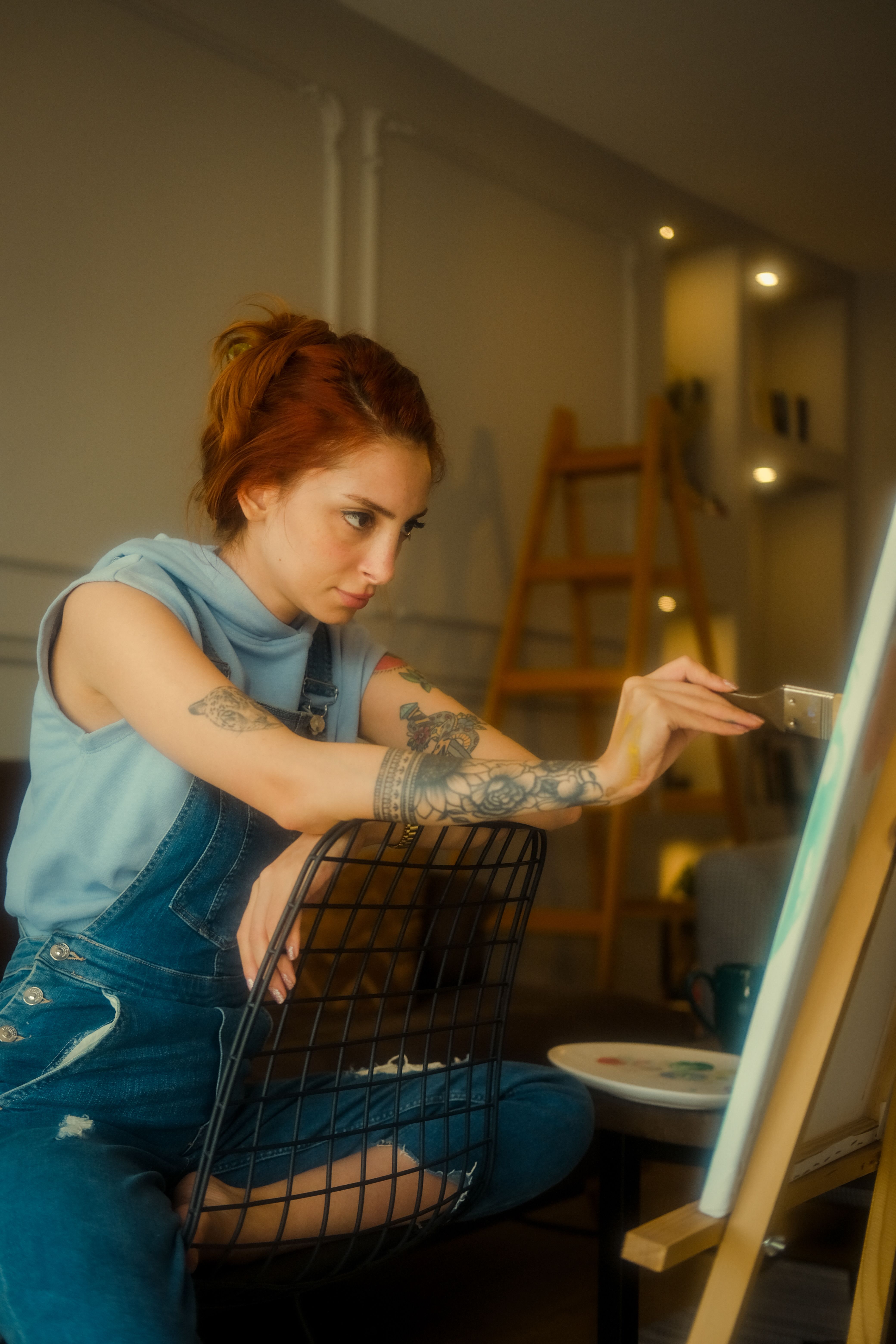 A redheaded woman artist concentrating on her painting, seated at an easel in a cozy indoor studio.