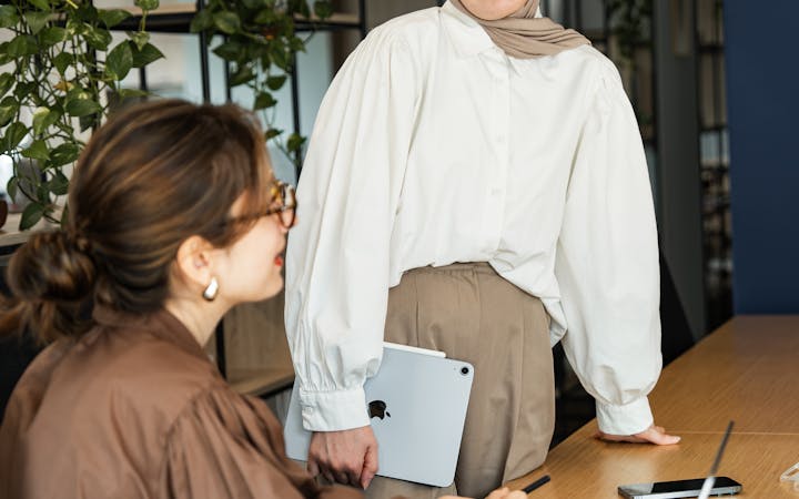 Two businesswomen in a modern office engaged in a work discussion, using laptops and notes.