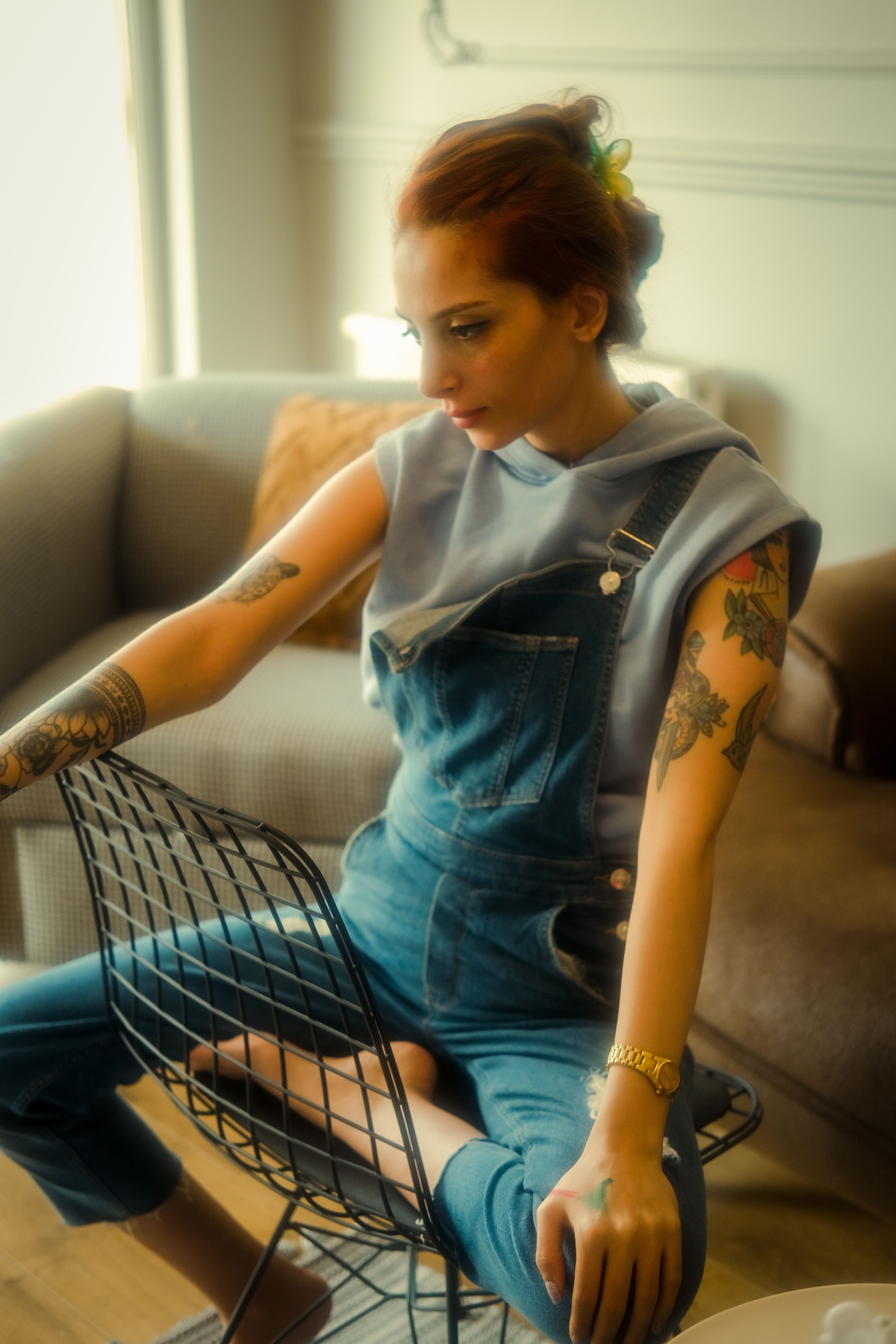 Fashionable redhead woman with tattoos sitting in a cozy room wearing denim overalls.