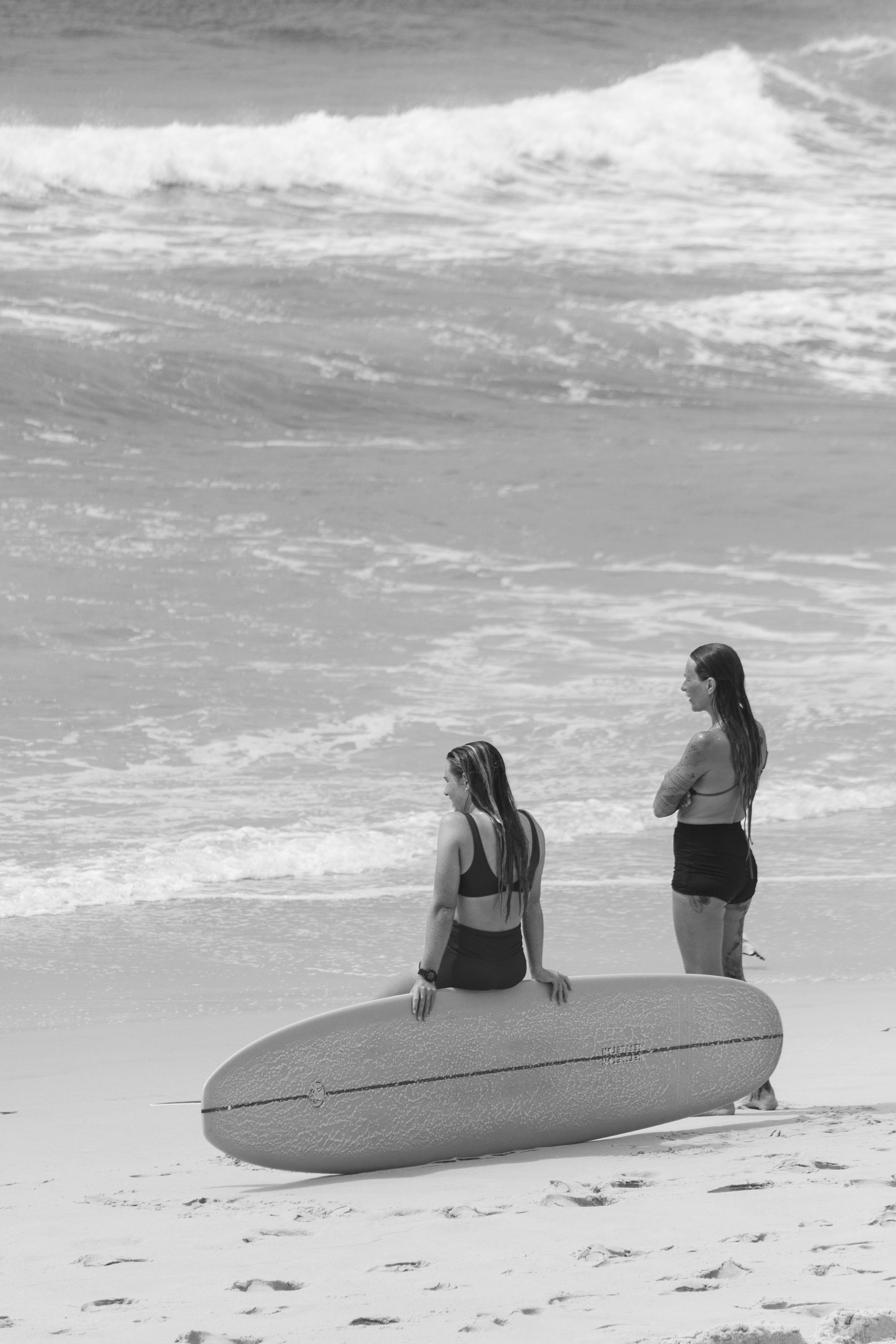 Two women with surfboards on Sydney beach, captured in black and white, evoke a serene coastal vibe.