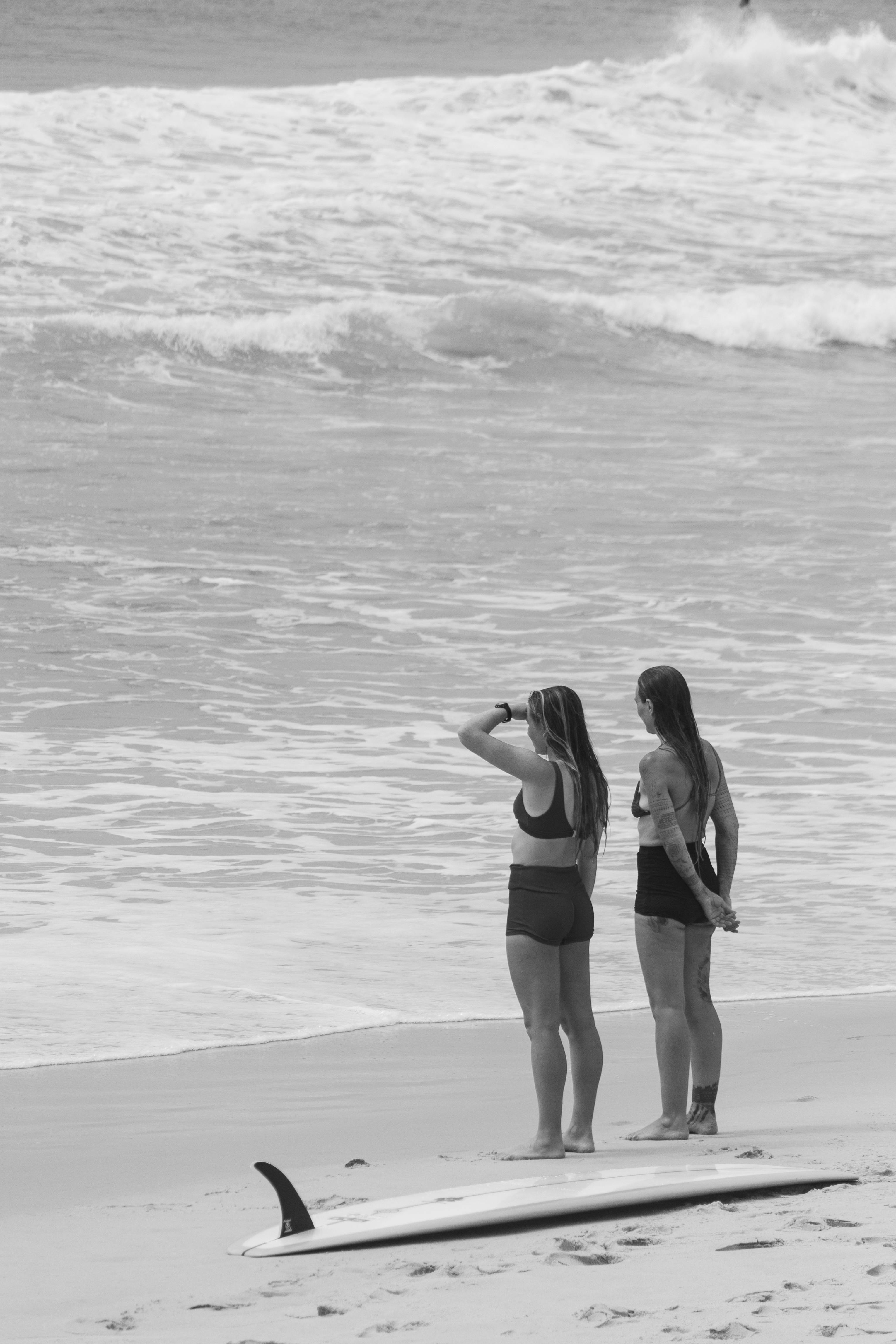 Two women in bikinis stand at Sydney beach, gazing at the sea near a surfboard.