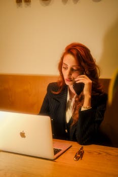 Red-haired woman in a café working on a laptop and talking on the phone.