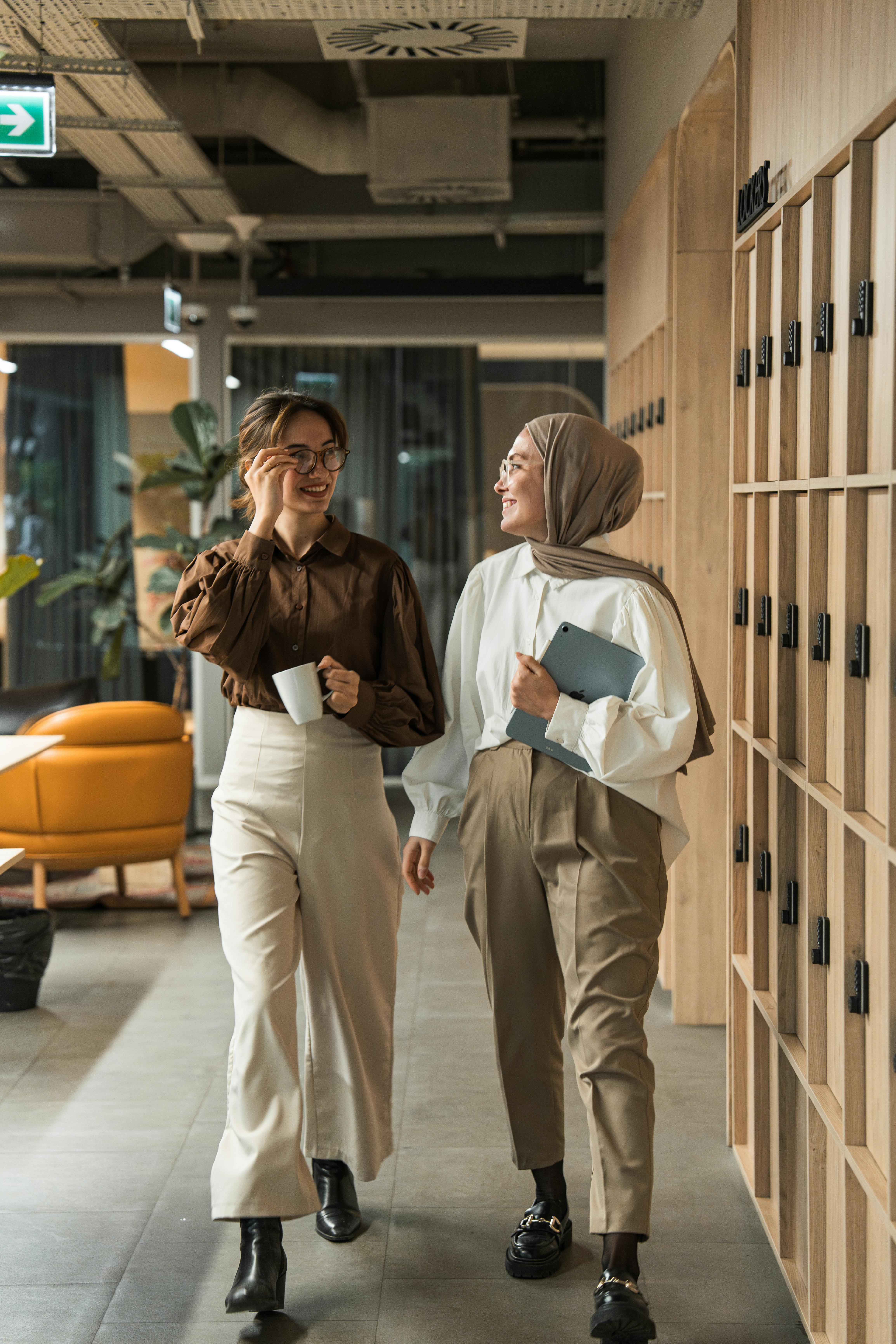 Two women in a modern office hallway, engaged in conversation and holding a tablet and mug.