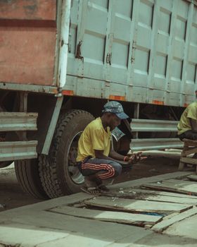 A man squatting beside a truck, utilizing tools in an urban outdoor setting.