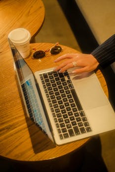 A hand typing on a laptop at a cafe table with a coffee cup and sunglasses.