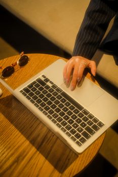 Close-up of a woman's hand using a laptop on a wooden table, beside sunglasses.