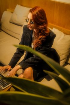 Redheaded woman in a cozy café working on a laptop, surrounded by soft lighting and plants.