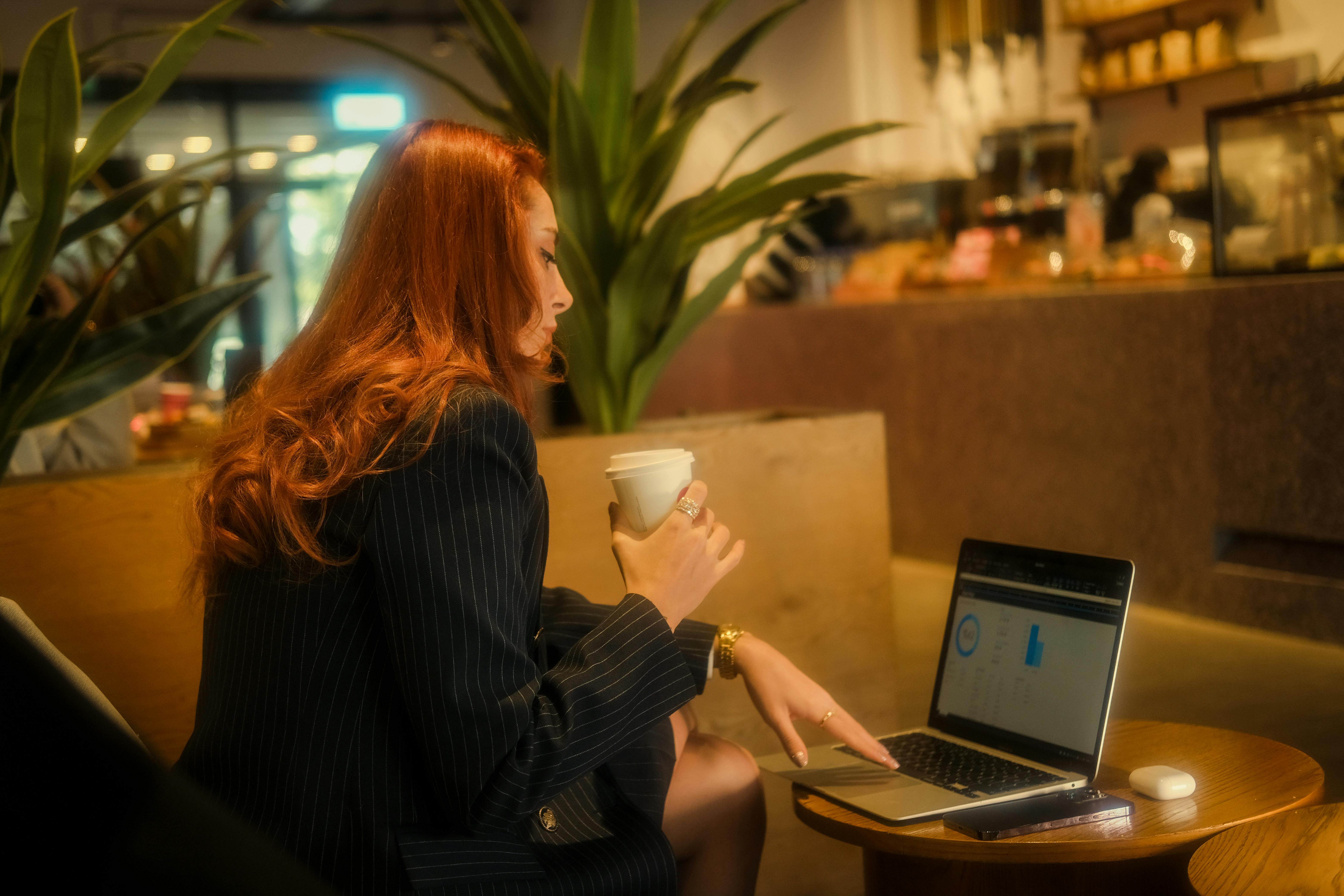 Redheaded businesswoman working on a laptop in a cozy café, holding a coffee.