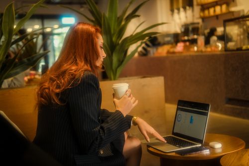 Free Redheaded businesswoman working on a laptop in a cozy café, holding a coffee. Stock Photo