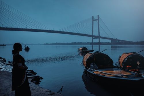Free Peaceful scene of a woman by boats and the iconic Howrah Bridge at dawn in Kolkata, India. Stock Photo