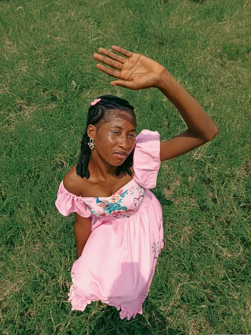 Free A young woman in a pink dress poses outdoors, shielding her eyes from the sun. Stock Photo