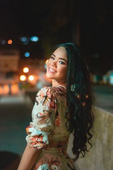 Portrait of a smiling woman with long hair in a floral dress standing outdoors at night.