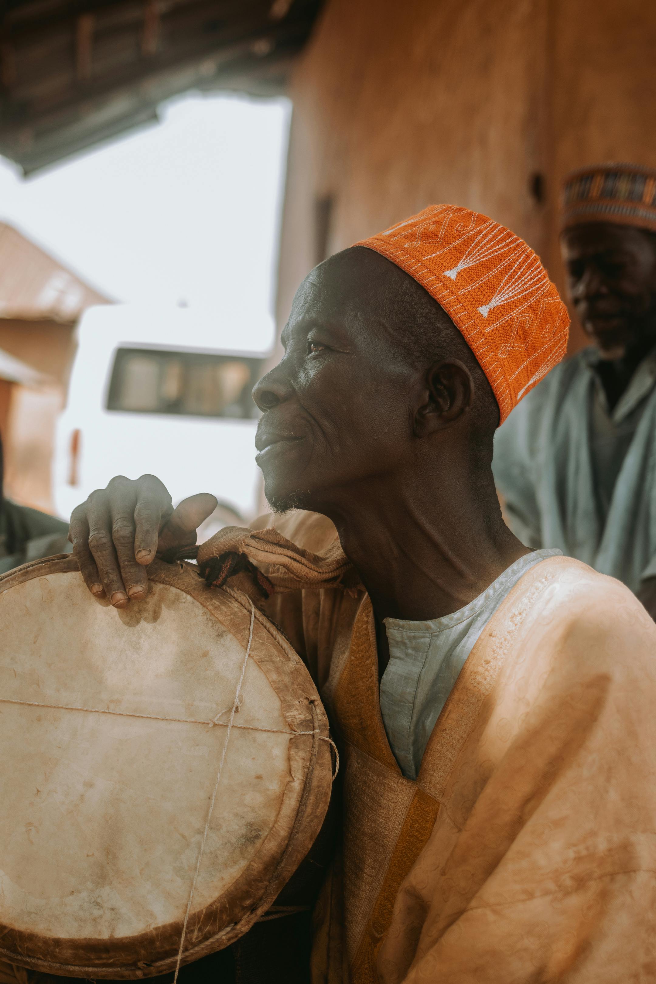 Elderly Nigerian man in traditional attire playing a drum outdoors in Essan.