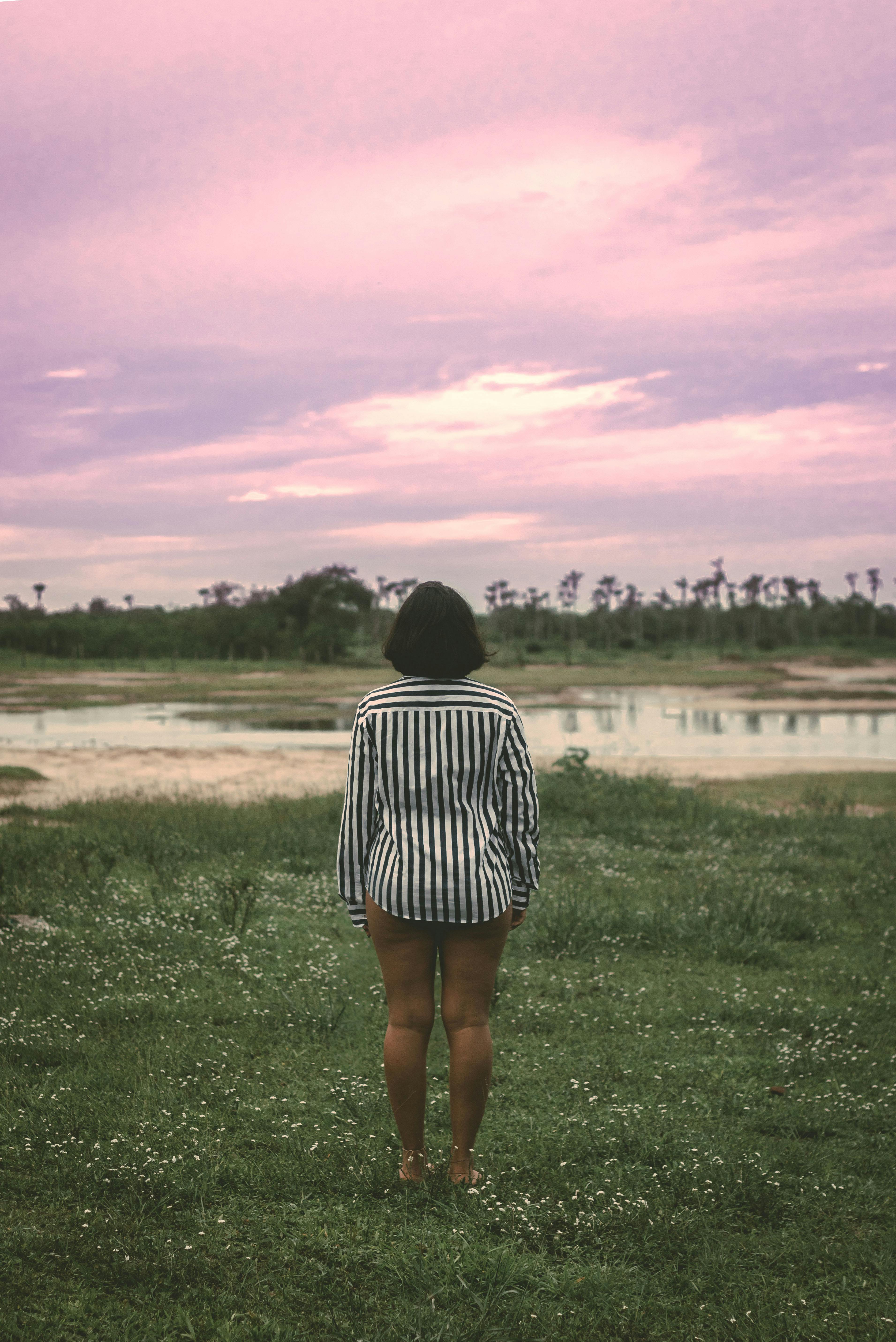 Back view of anonymous female traveler in casual clothes standing on grassy meadow near pond and admiring amazing sunset in countryside