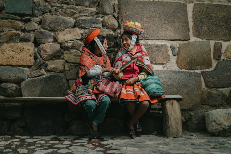 Peruvian Girls Wearing Traditional Clothing, Sitting On A Bench By A Wall