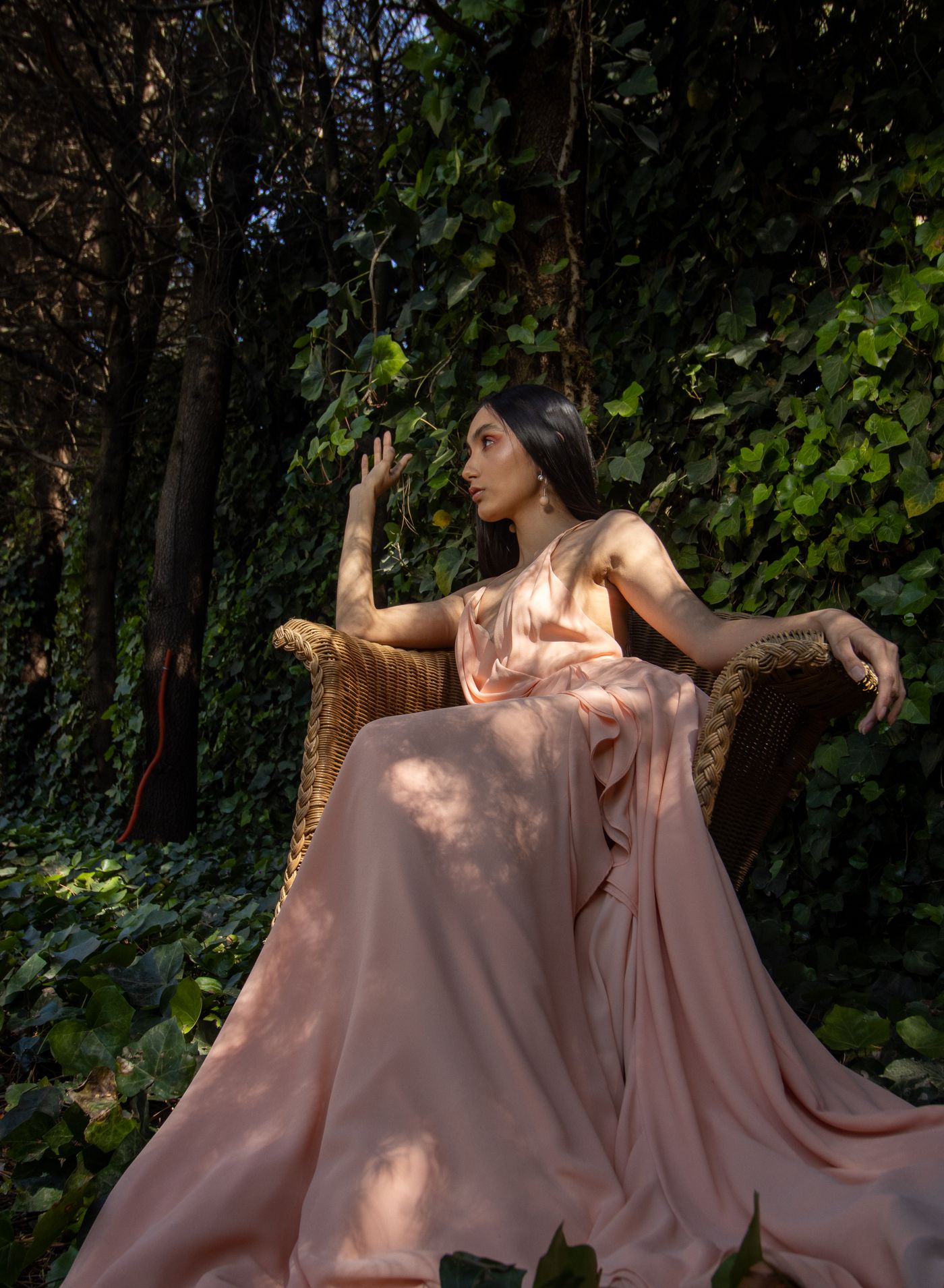 A model in a pink evening dress seated in a wicker chair among densely growing ivy