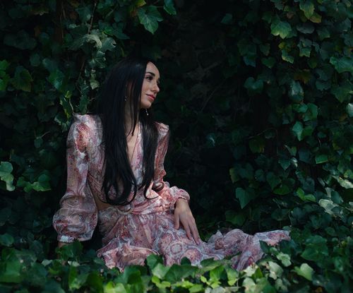 Free Elegant woman in floral dress seated in lush green ivy outdoors. Stock Photo