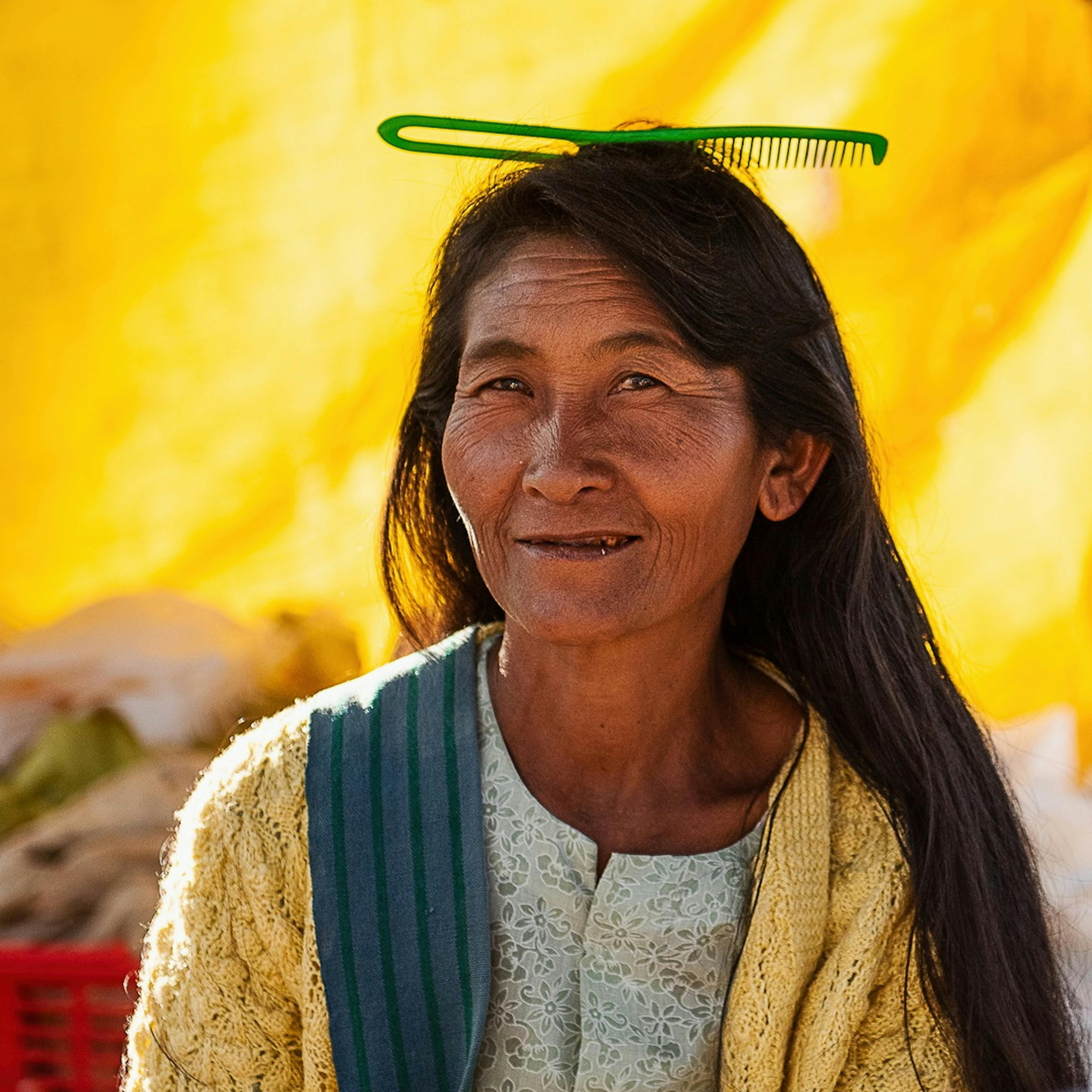 An elderly woman with long hair wearing a comb, smiling warmly against a vibrant yellow background.
