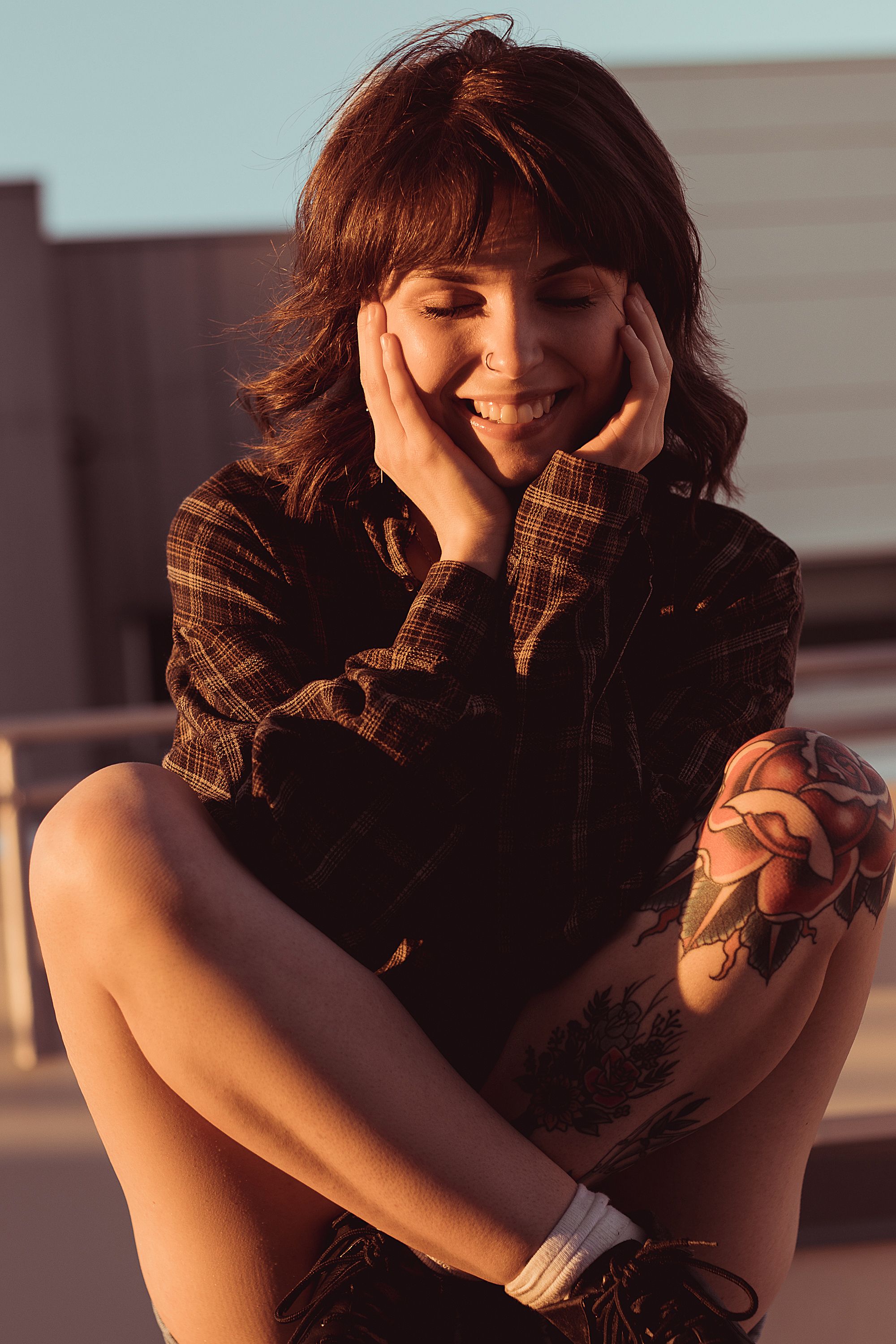 Portrait of a young woman with tattoos smiling warmly in the sunset, seated outdoors.