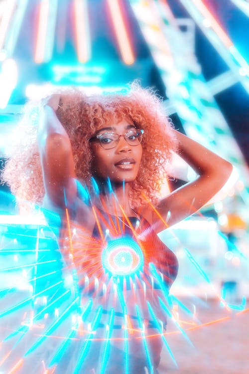 A woman with curly hair and glasses is standing in front of a neon light