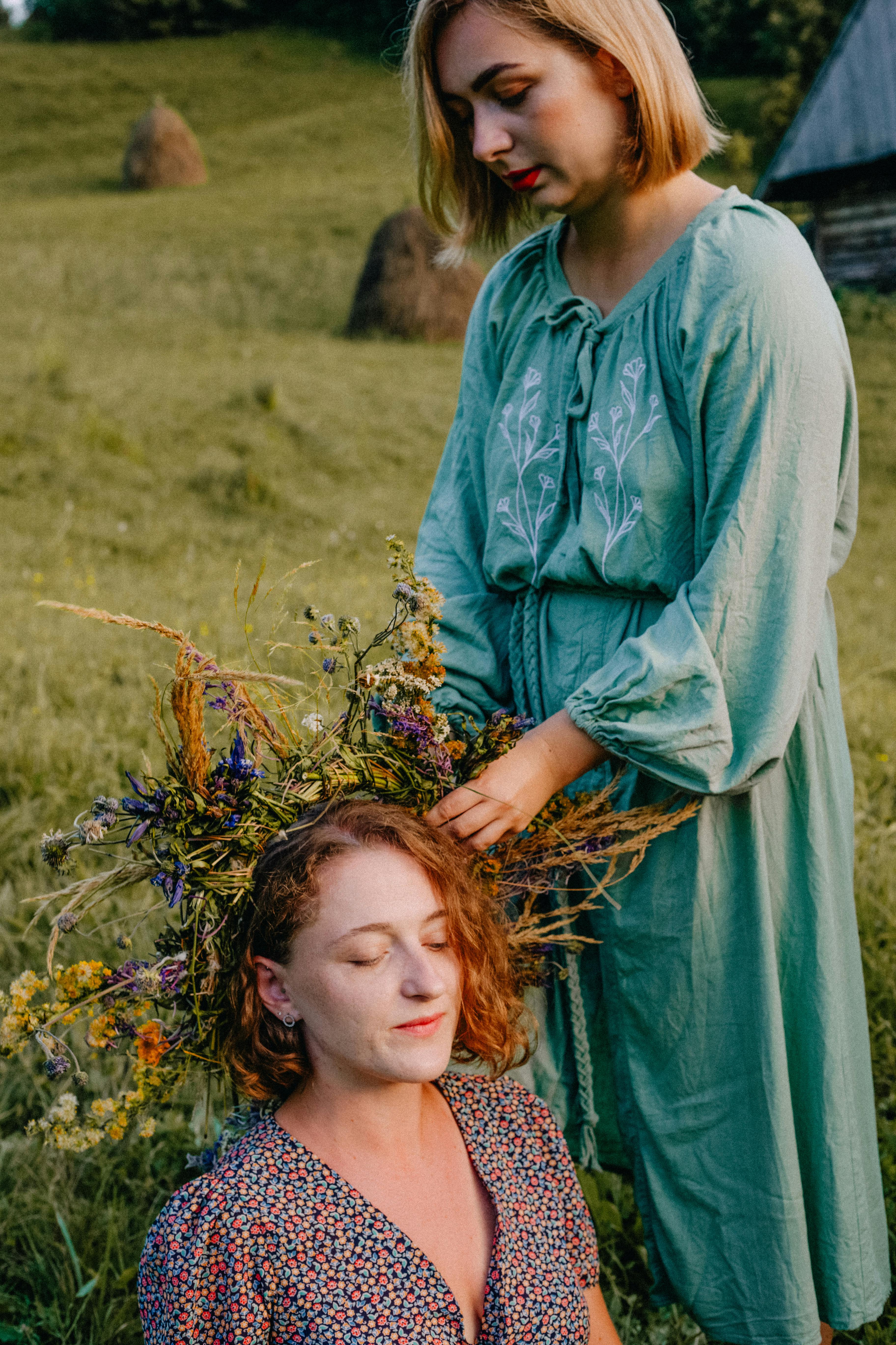 Two women in floral dresses enjoy nature, creating a flower wreath on a sunny day.