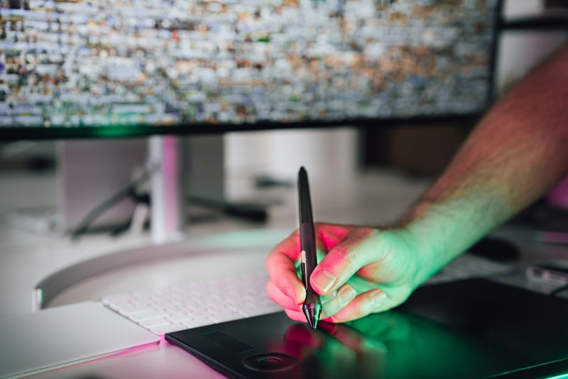 Mano con un lápiz óptico sobre una tableta gráfica bajo un monitor, con luz de borde verde y rosa