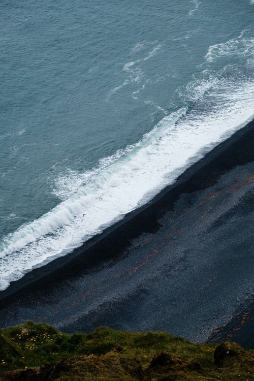 Free A dramatic aerial shot of waves crashing on a black sand beach with lush greenery. Stock Photo