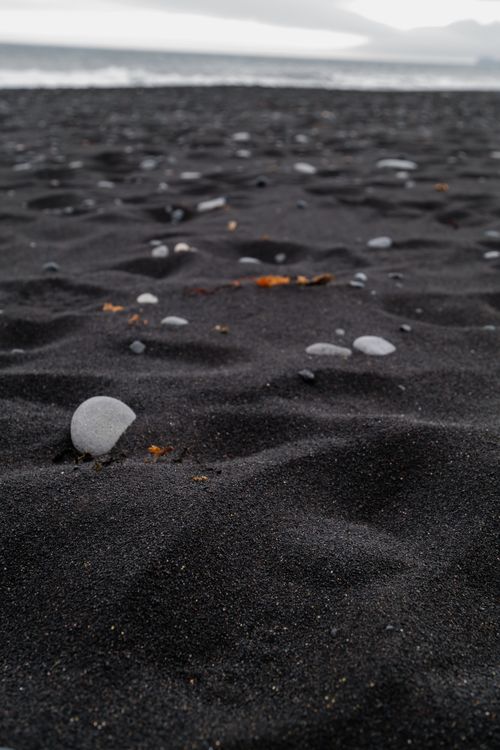 Free Close-up view of a black sand beach with scattered smooth pebbles and ocean waves in the background. Stock Photo