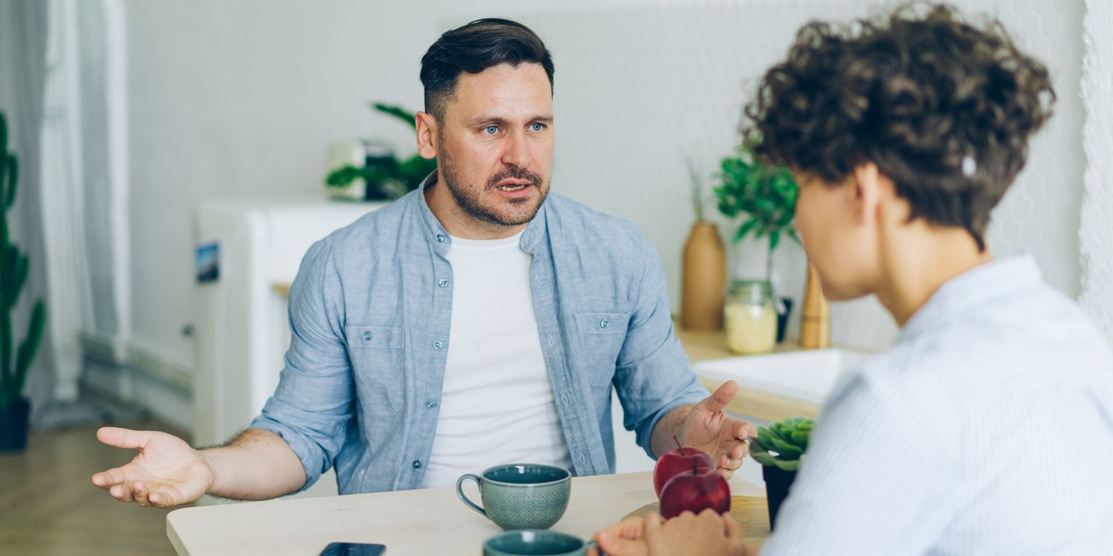 Two adults engage in a conversation at a kitchen table with coffee cups.
