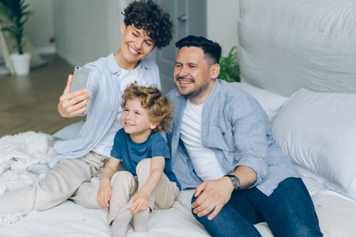 Free Smiling parents and child taking a selfie on a bed, capturing a joyful moment together. Stock Photo