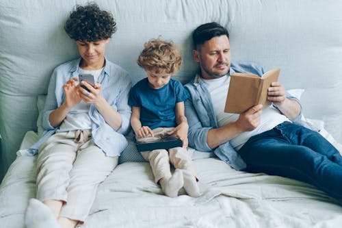 A Family with a Little Son Sitting on a Bed, Mother Looking at Her Smartphone and Father Reading a Book