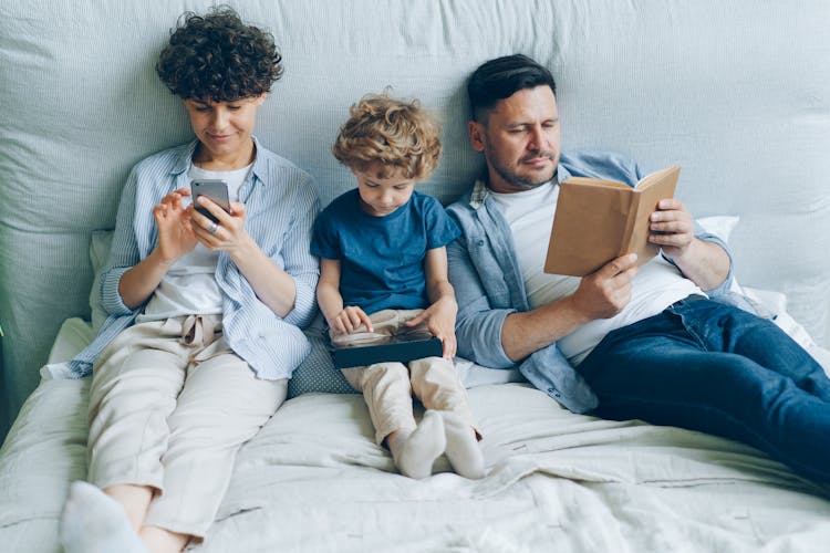 A Family With A Little Son Sitting On A Bed, Mother Looking At Her Smartphone And Father Reading A Book