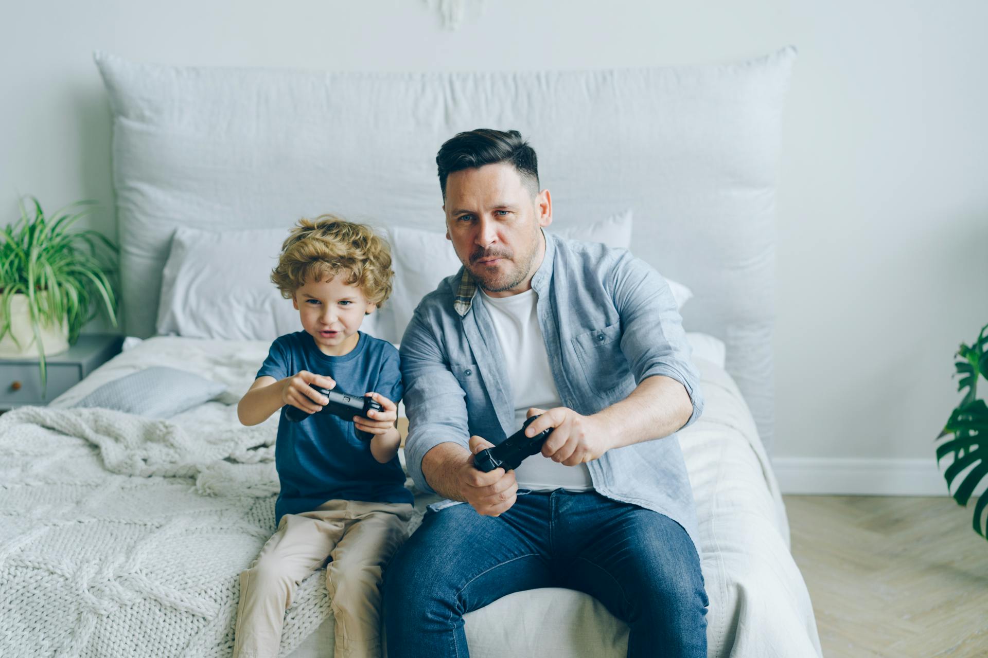 Man and His Little Son Sitting on a Bed and Playing a Video Game