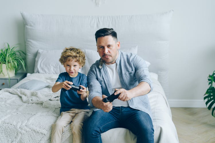 Man And His Little Son Sitting On A Bed And Playing A Video Game 