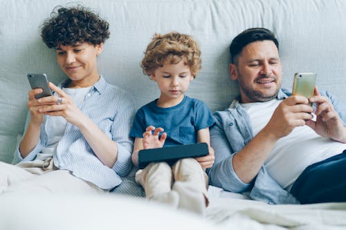 A Family with a Little Son Sitting on a Bed, Parents Looking at Their Smartphones and Their Son Playing a Game