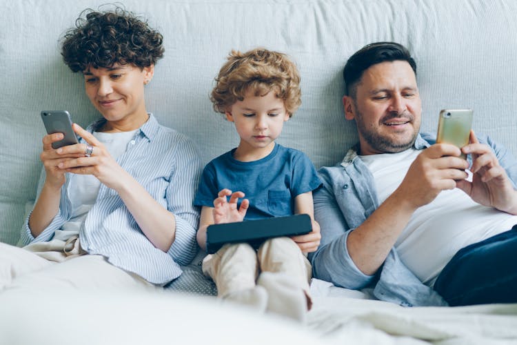 A Family With A Little Son Sitting On A Bed, Parents Looking At Their Smartphones And Their Son Playing A Game