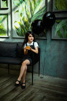 A young girl in a white shirt and dress holds a birthday cake next to black balloons indoors.