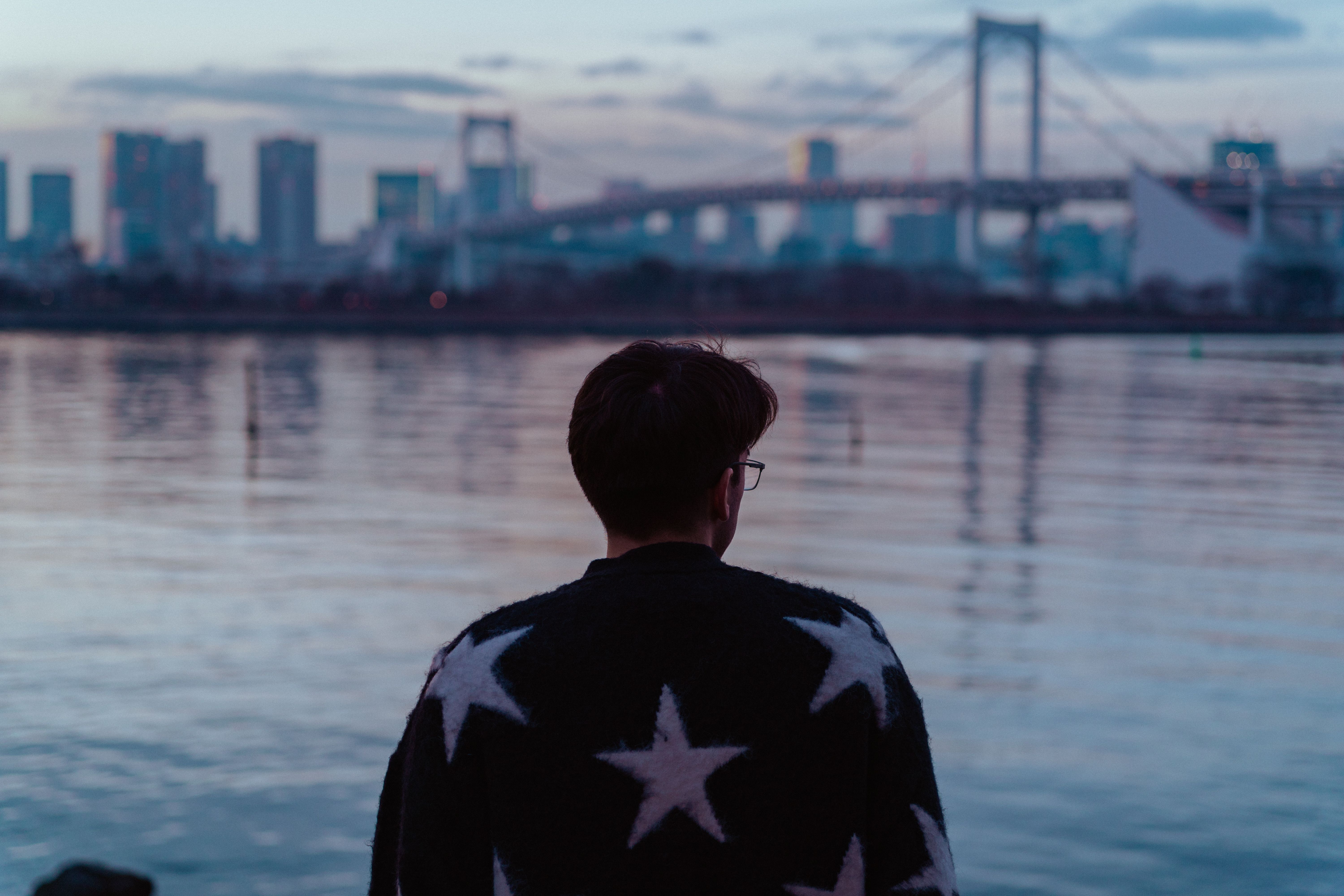 A man gazes at the Tokyo skyline and Rainbow Bridge at twilight, creating a reflective urban scene.