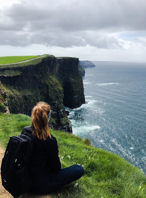 Woman Sitting on Sea Cliff Under Cloudy Sky