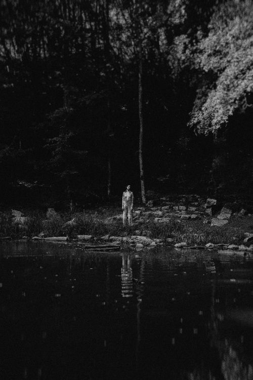 Free A solitary figure standing by a lake reflecting trees and a forest, in an eerie nighttime setting. Stock Photo