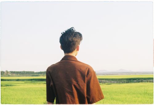 Free Rear view of a man in a brown shirt standing in a lush green field under a clear sky. Stock Photo