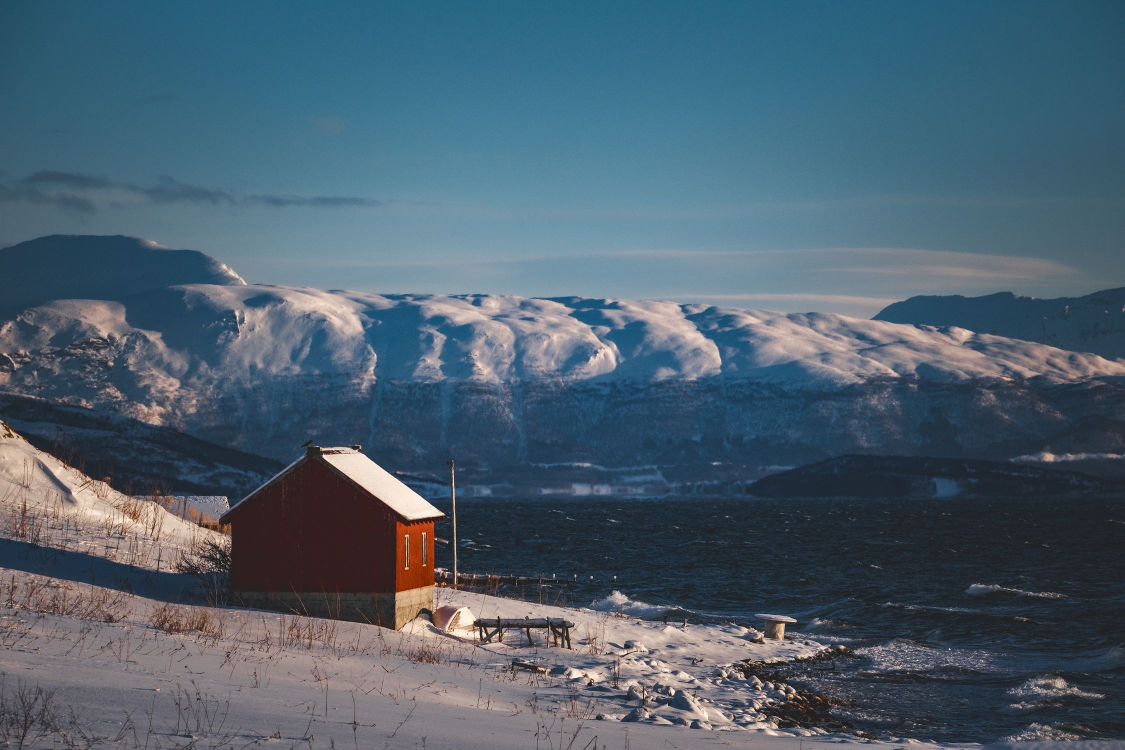 A picturesque snowy cabin by a lake surrounded by majestic mountains in winter.