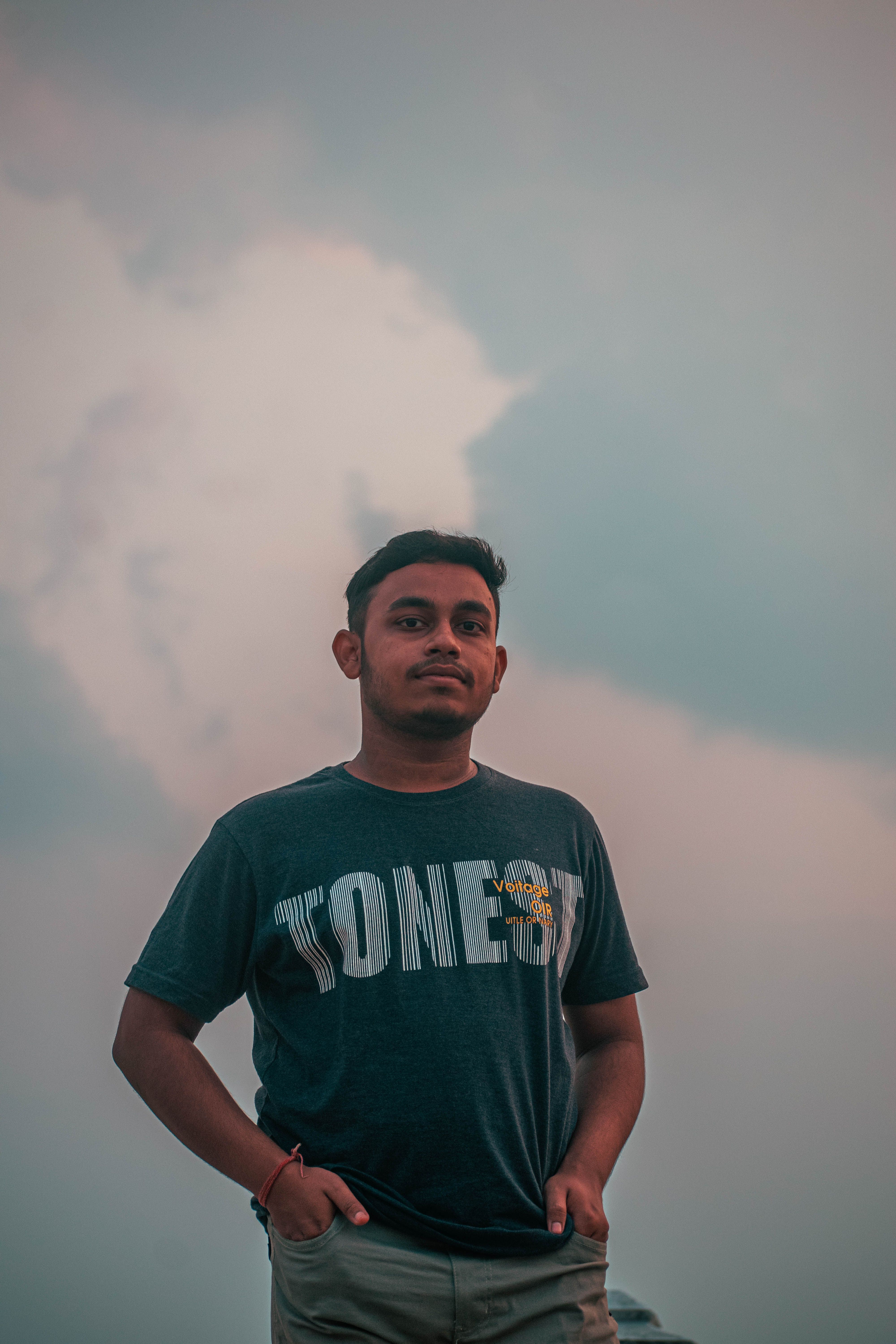 A young man stands outdoors at sunset with hands in pockets, showcasing casual fashion.