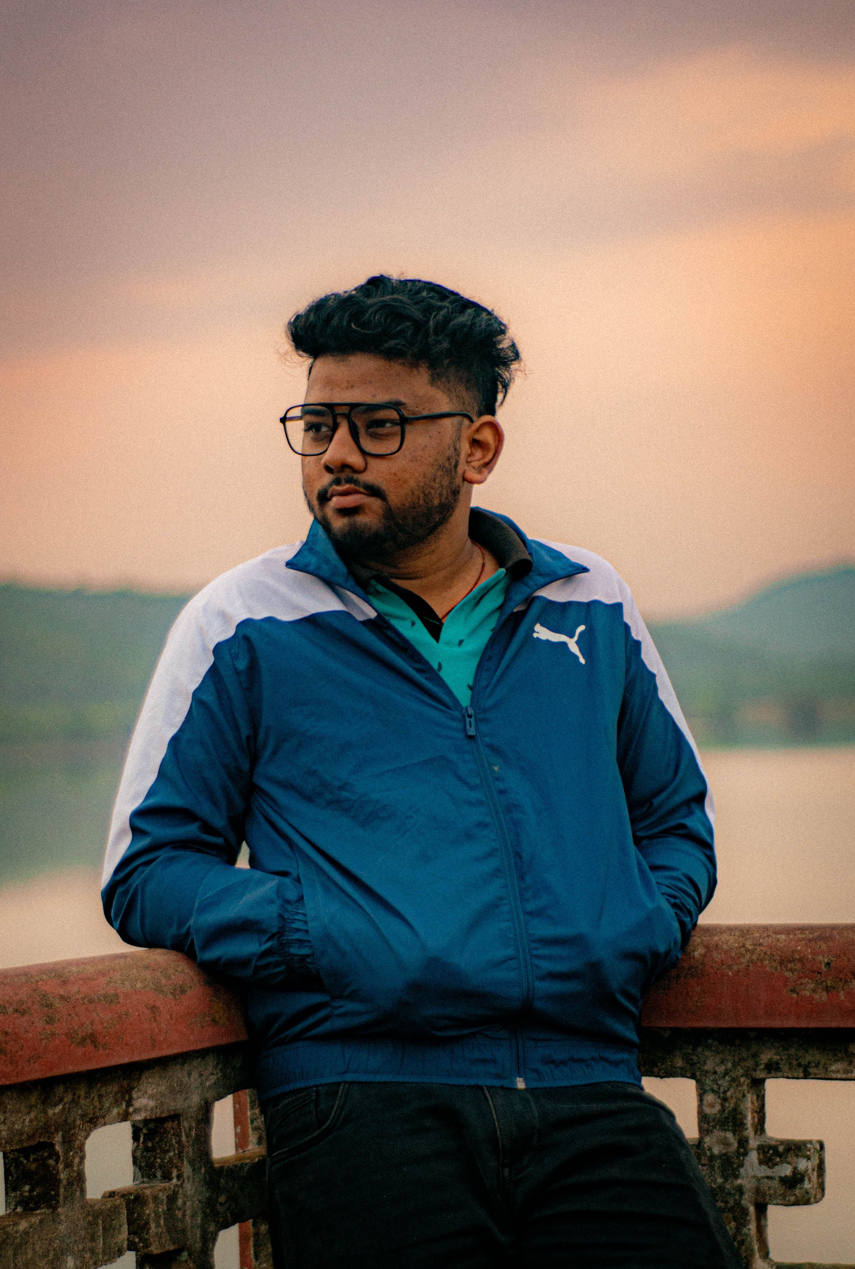Portrait of a man with eyeglasses in a blue jacket, leaning on a railing at sunset.