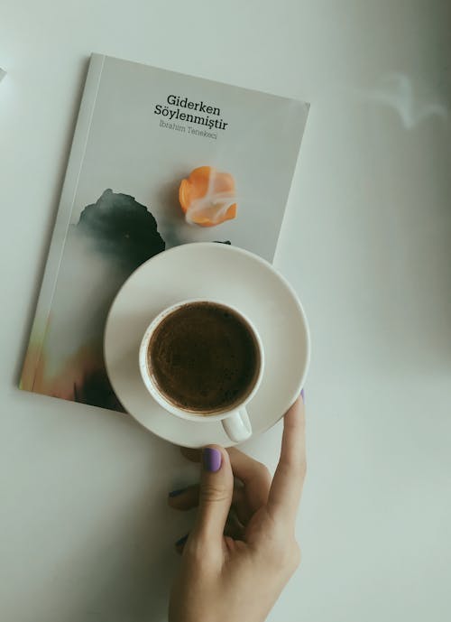 Female Hand Holding a Cup of Coffee Standing Next to a Book