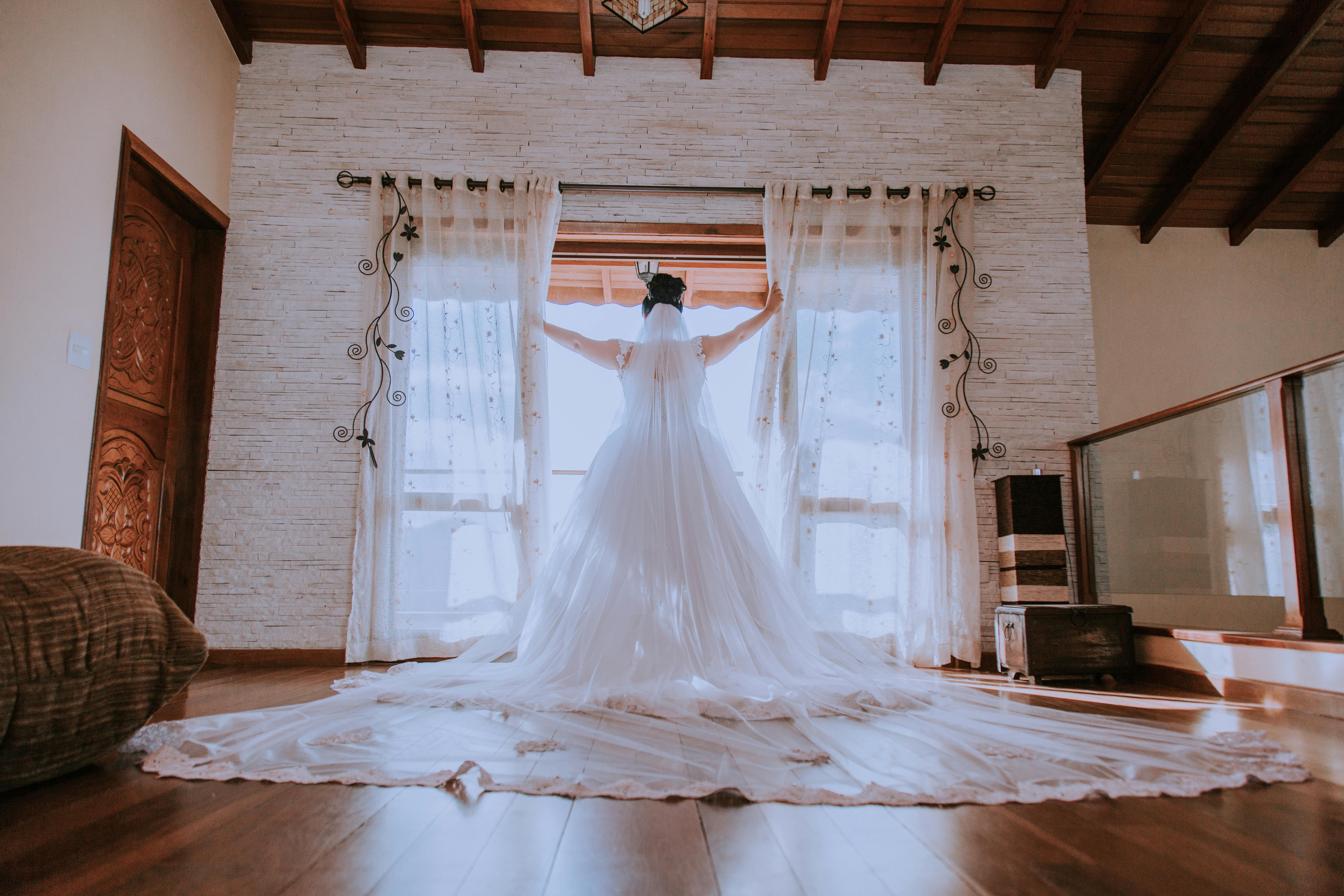 Bride in a flowing gown stands before a sunlit window in an elegant room.