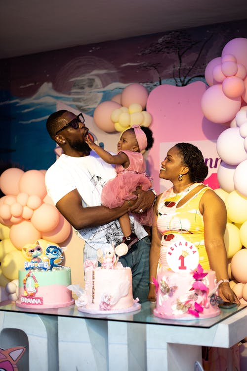 Parents with a Baby Girl Smiling and Standing in Front of Birthday Cakes with Decorations Around
