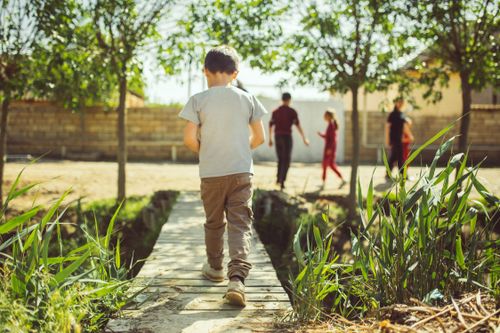 Free Young boy walks on a footbridge in a park during summer, surrounded by greenery and friends in the background. Stock Photo
