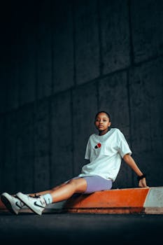 Young African woman in casual attire sitting on a barrier in a dimly lit urban setting.