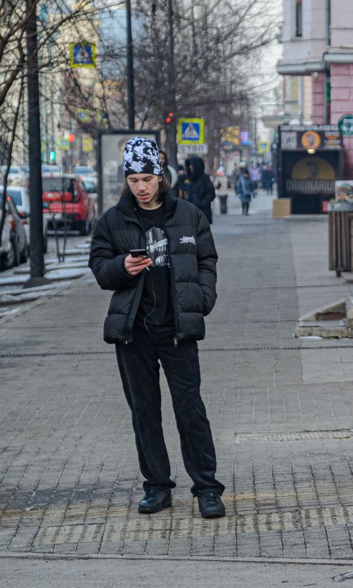 Free Casual young man standing on a city sidewalk in winter attire, engrossed in his smartphone. Stock Photo